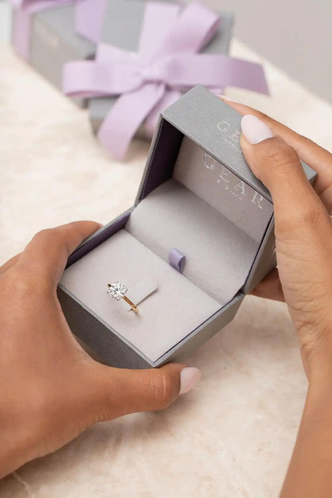 Delicate engagement ring with a single stone in a gray box with a lavender bow.