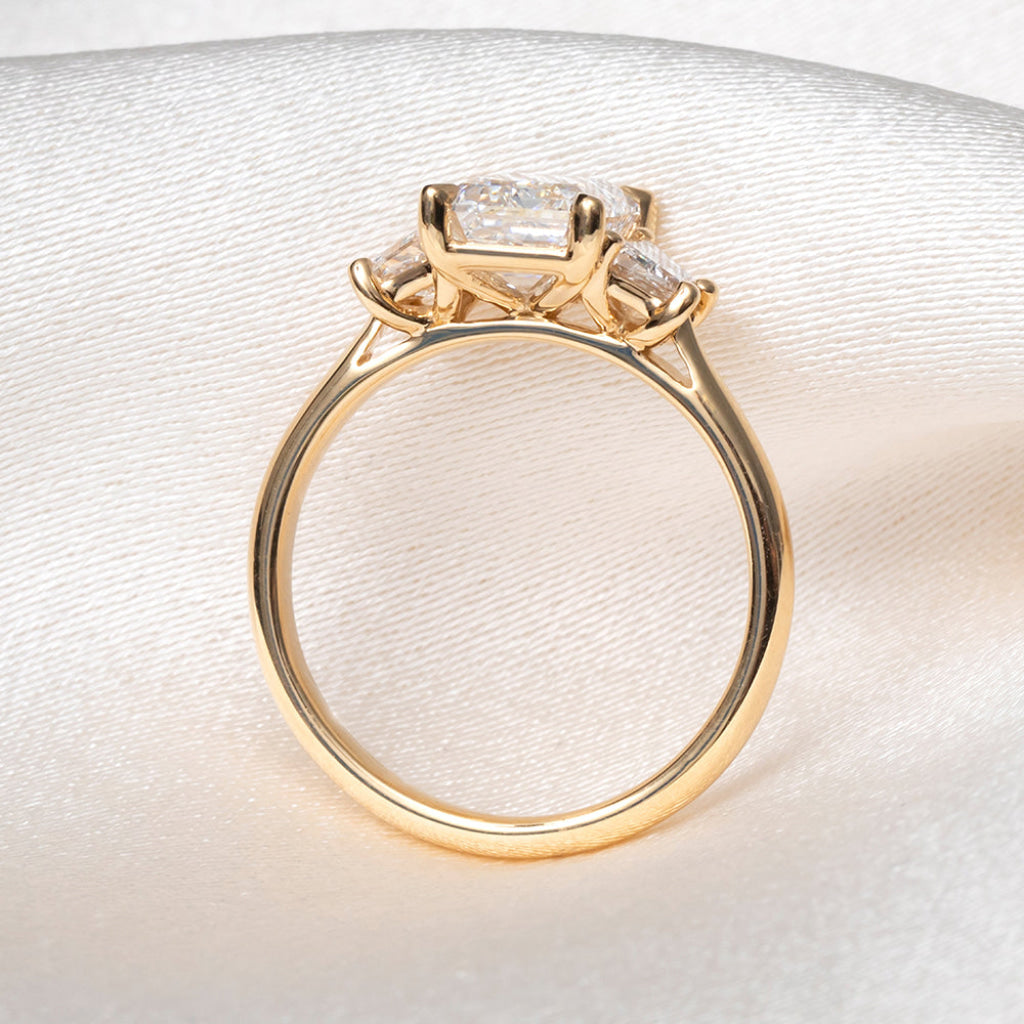 A yellow gold ring with a large rectangular clear gemstone and smaller side stones, photographed on a soft white satin-like fabric background.