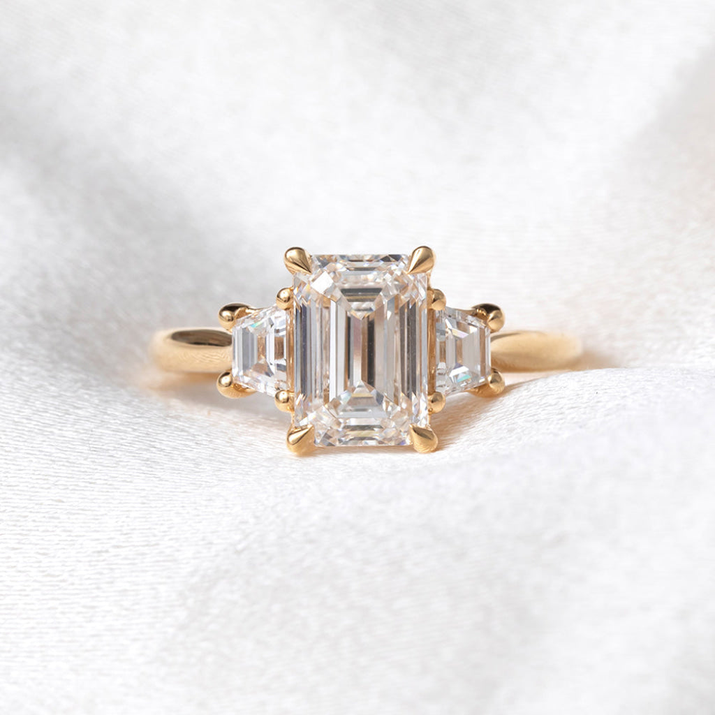 A yellow gold three-stone ring featuring a large emerald-cut center stone and two smaller emerald-cut side stones, displayed on a soft white textured fabric background.