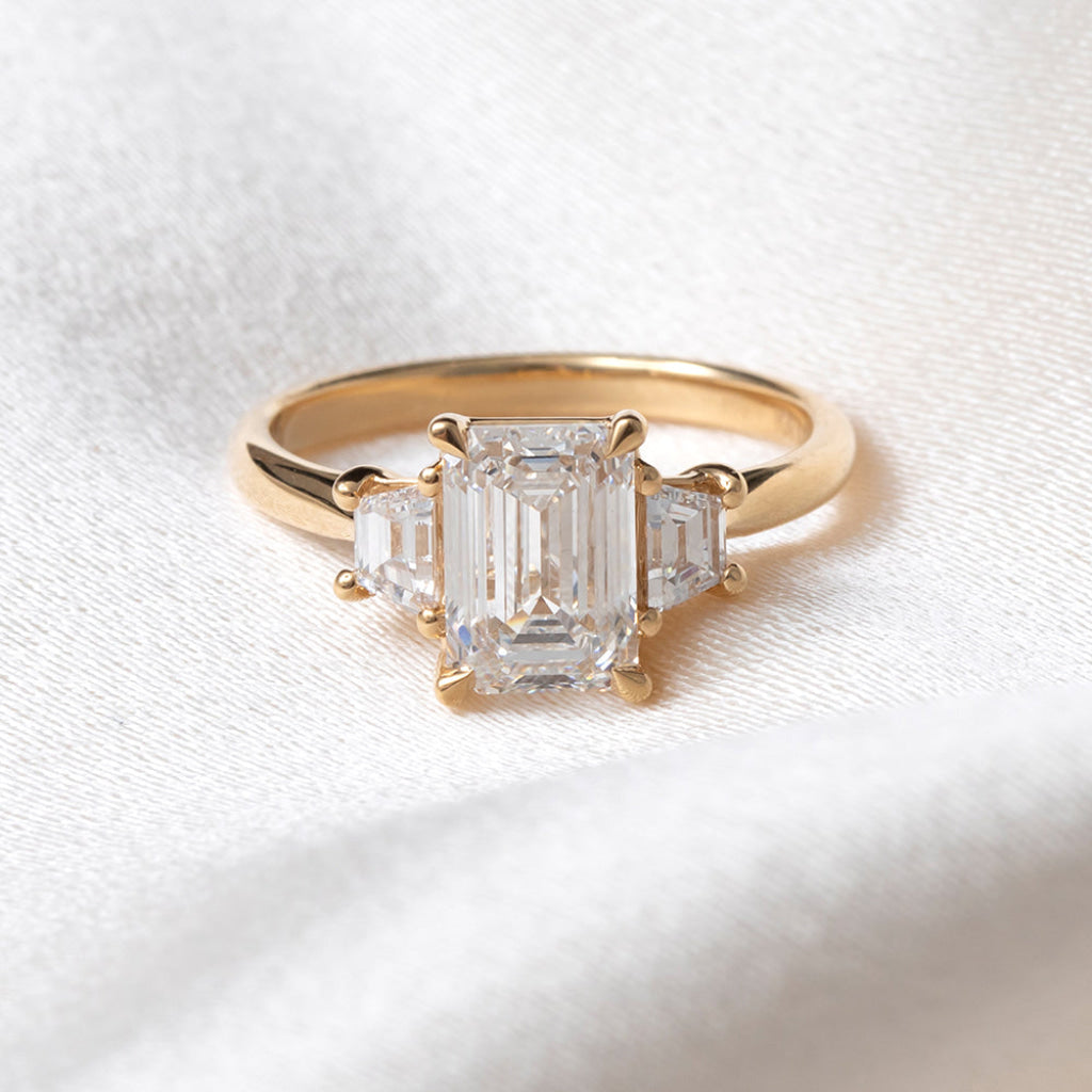 A yellow gold three-stone ring featuring a large emerald-cut center stone and two smaller baguette-cut side stones, displayed on a textured white fabric background.