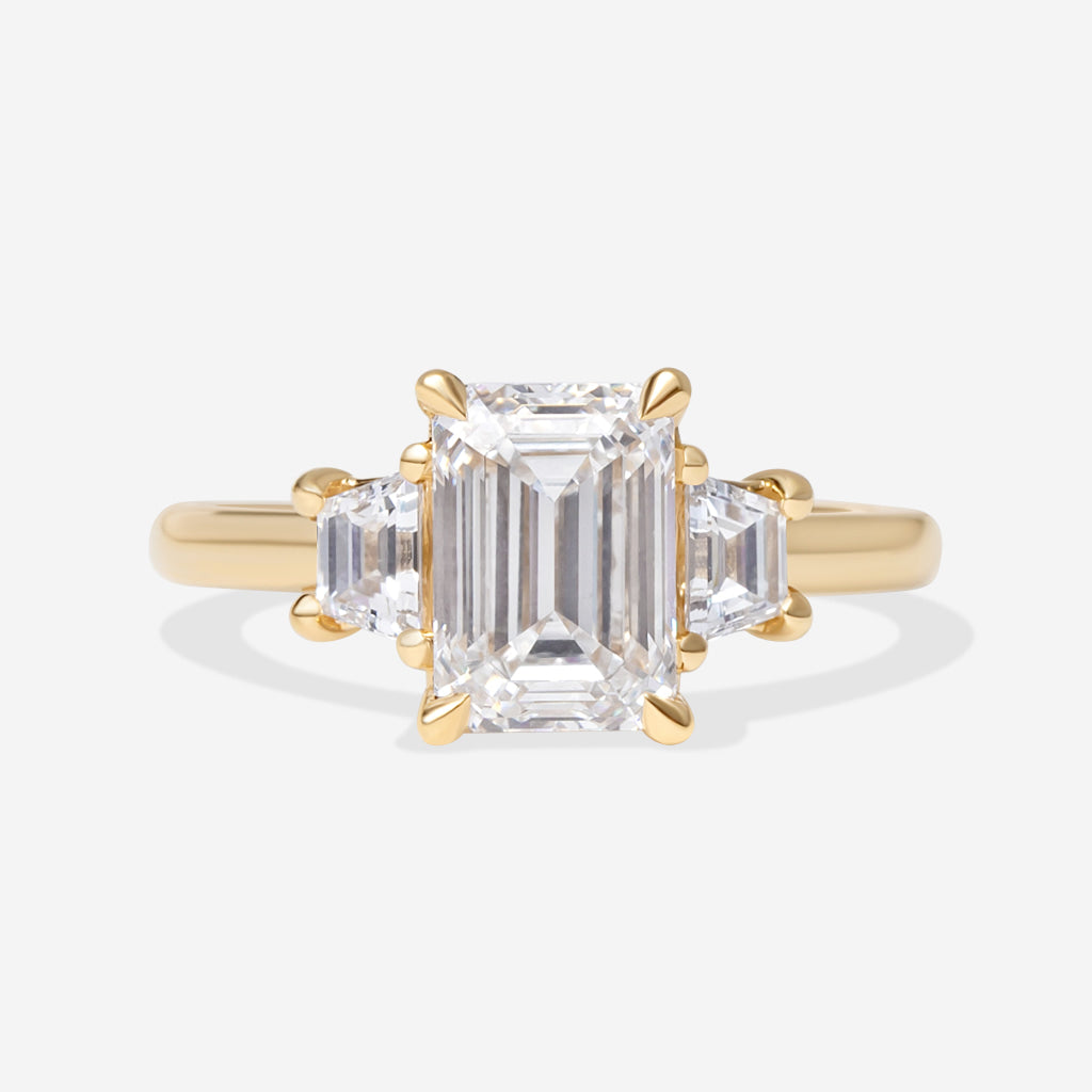 A yellow gold three-stone ring featuring a large emerald-cut center diamond flanked by two smaller baguette-cut side diamonds, set against a clean white background.