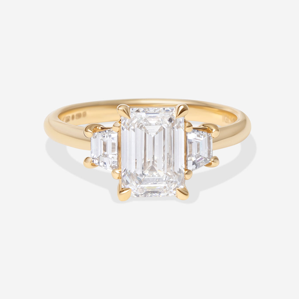 A yellow gold three-stone ring featuring a large emerald-cut center diamond flanked by two smaller round diamonds, set against a clean white background.