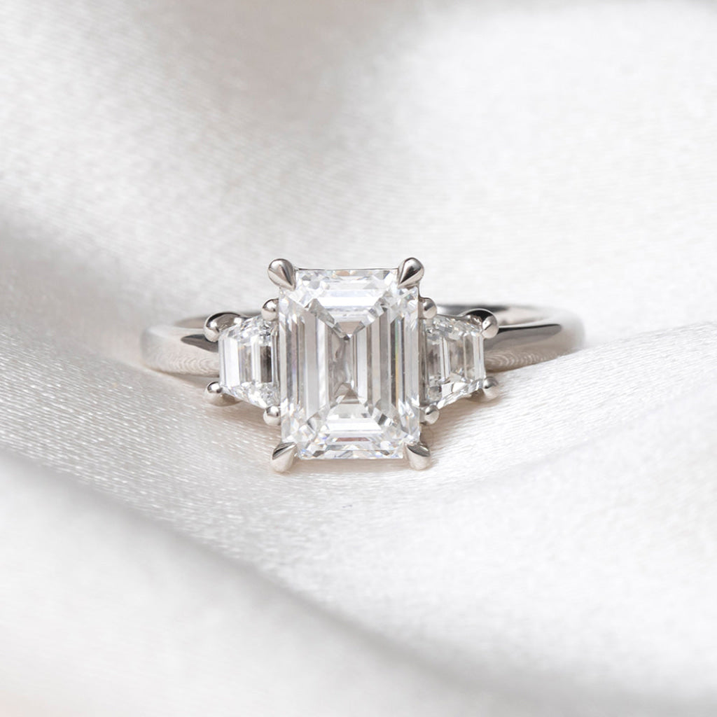 An emerald-cut diamond ring with baguette side stones on a platinum band, resting on a textured white fabric background.