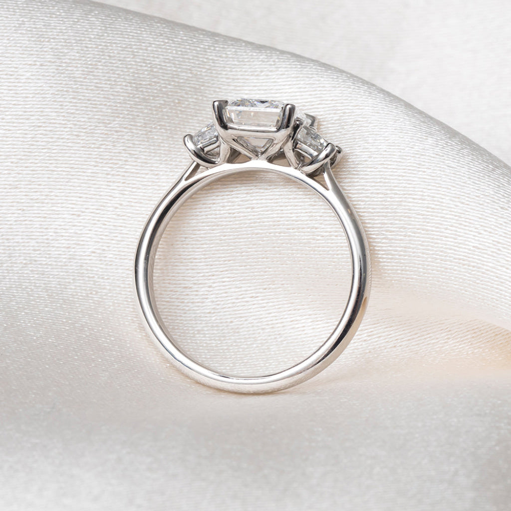 A platinum three-stone ring with a rectangular center stone and smaller side stones, displayed on a textured white satin fabric background.