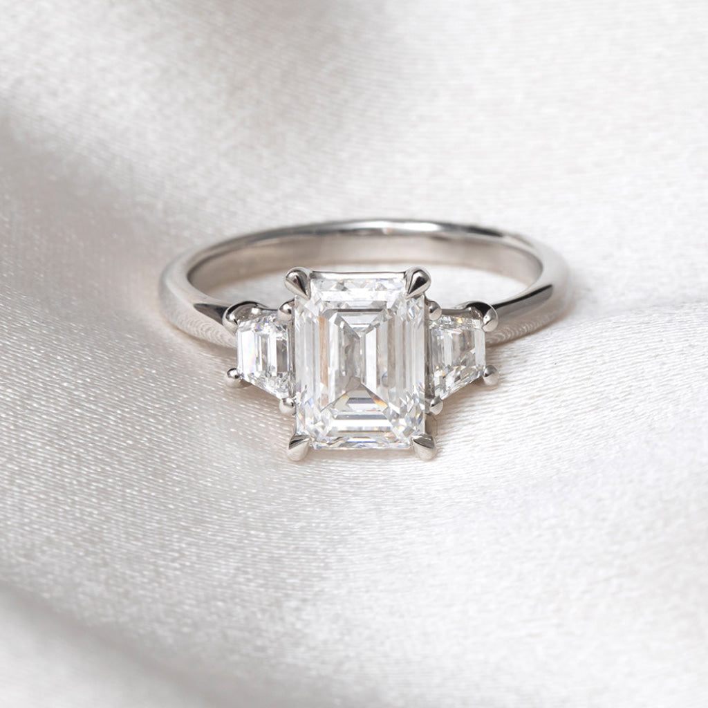 An emerald-cut diamond ring with a three-stone setting on a polished platinum band, resting on a textured white fabric background.