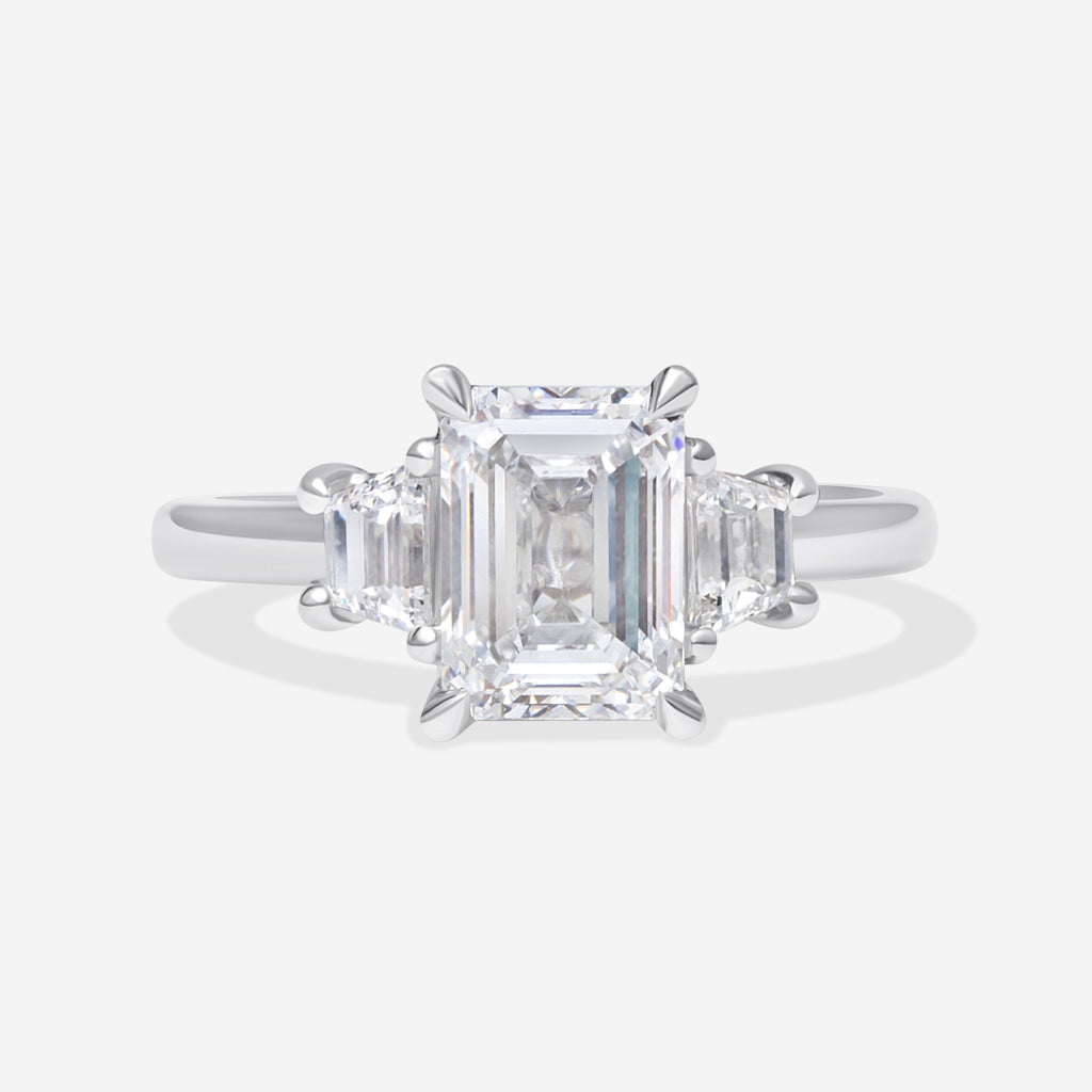 A platinum three-stone ring featuring a large emerald-cut center diamond flanked by two smaller trapezoid diamonds, set against a clean white background.