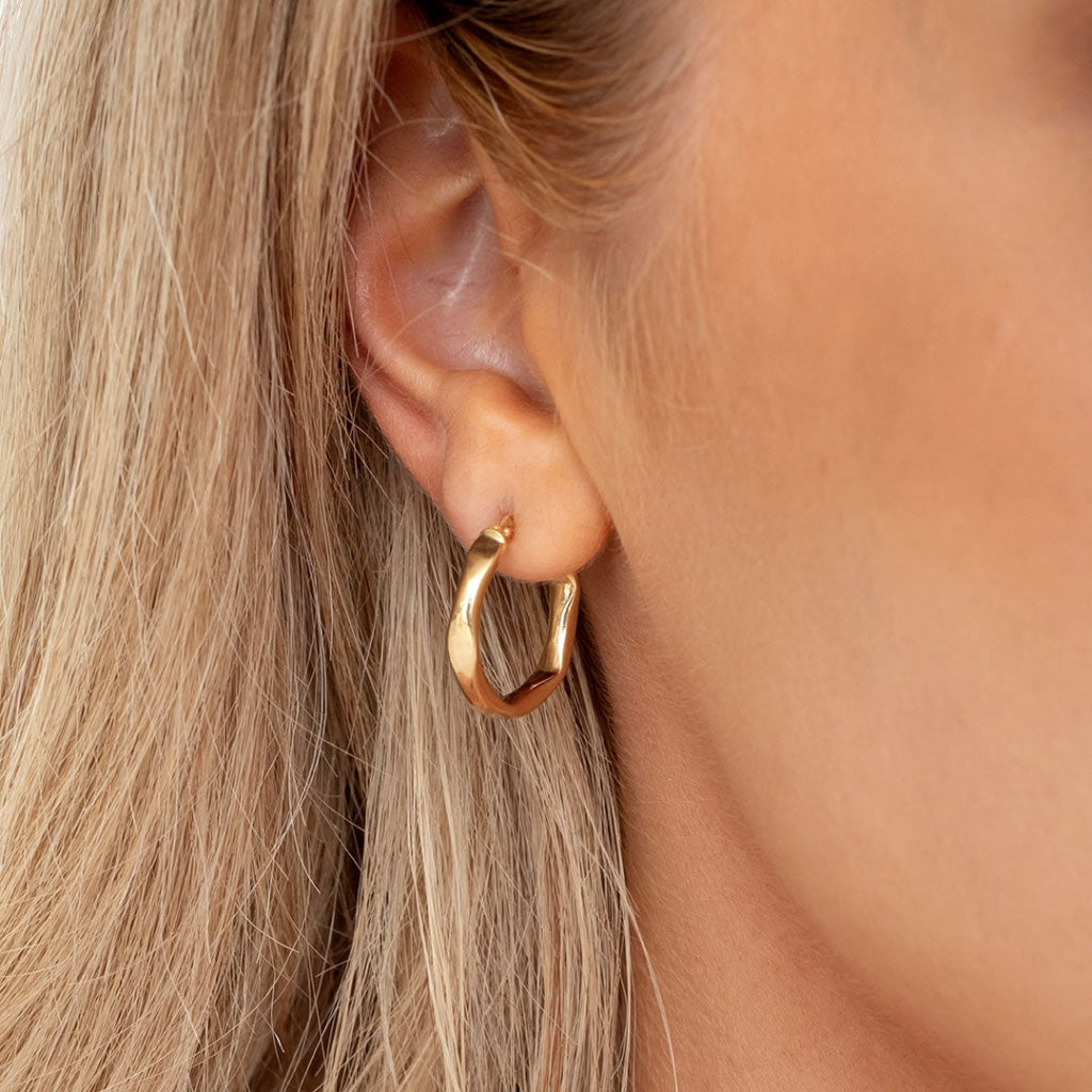 Side view of a woman's ear adorned with a sculpted gold hoop earring, its unique shape and gleaming surface highlighted against her blonde hair
