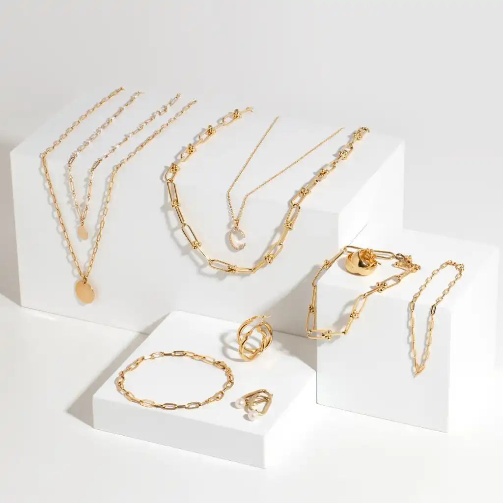 Collection of gold-toned chain necklaces, bracelets and rings displayed on white platforms.