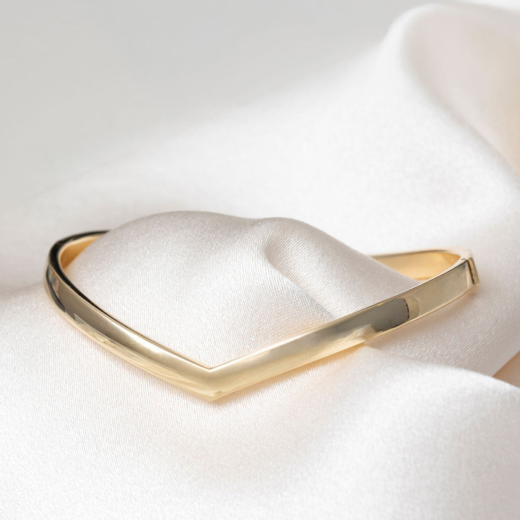A polished gold bangle bracelet with a smooth, rounded triangular design, displayed on a soft white satin fabric background.