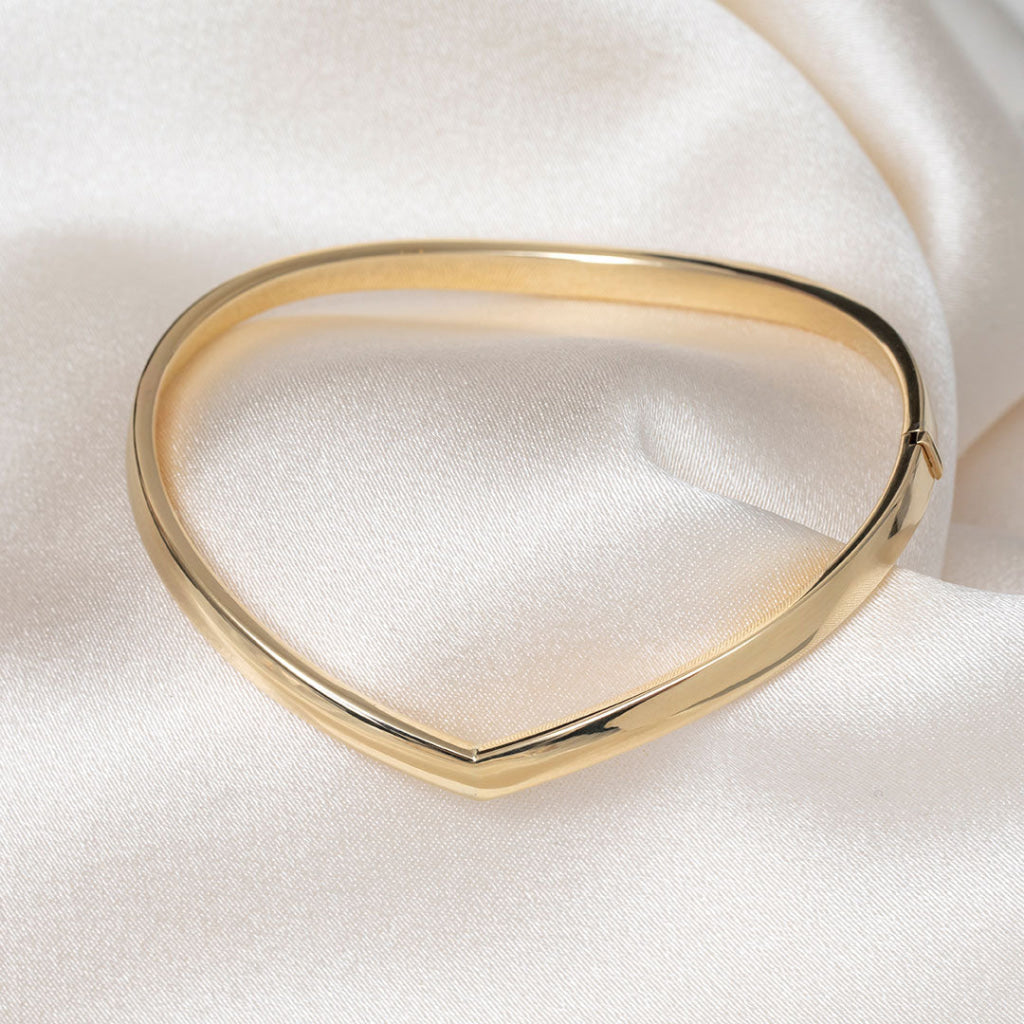 A polished gold bangle bracelet with a smooth, rounded triangular design, displayed on a soft white satin fabric background.