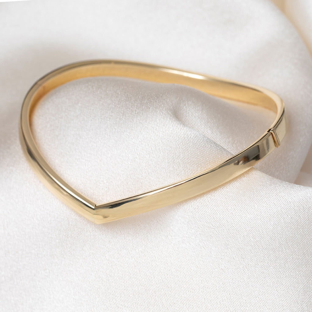 A polished gold bangle bracelet with a smooth, rounded triangular design, displayed on a soft white satin fabric background.