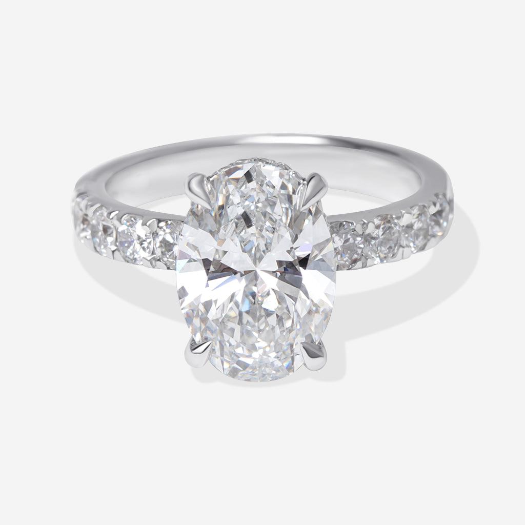 An oval-cut lab-grown diamond engagement ring with a platinum band featuring pavé-set round diamonds, shown on a light gray background.