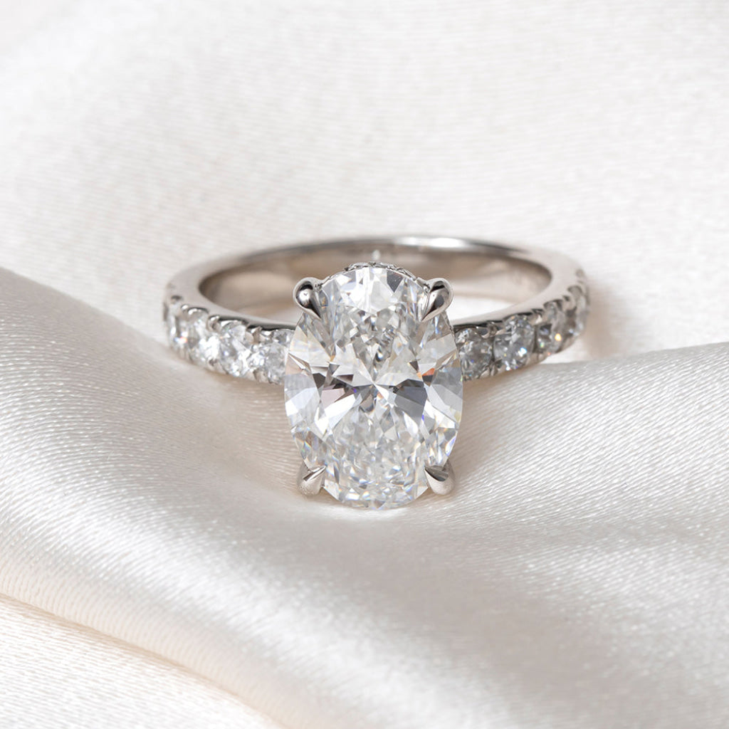 An oval-cut  lab grown diamond engagement ring with a platinum band set with round brilliant cut diamonds, resting on a soft white satin fabric.