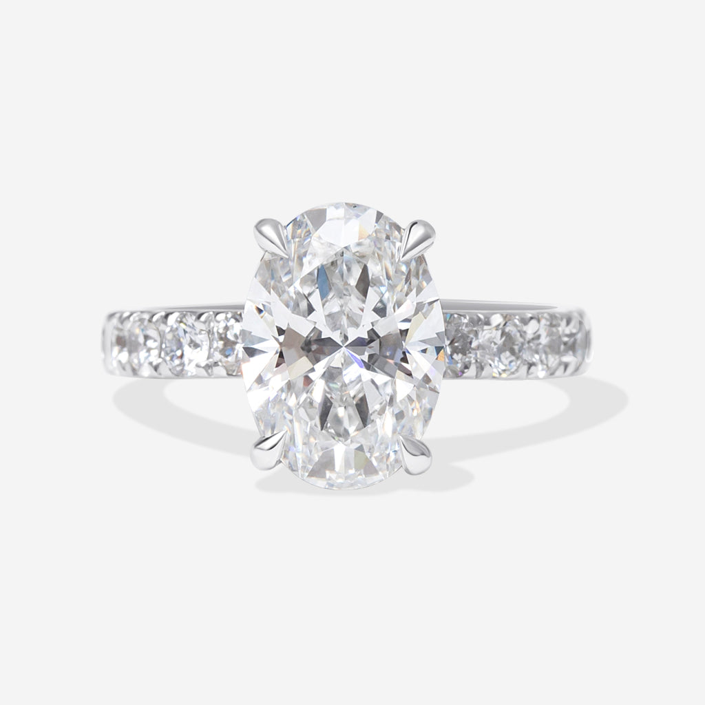 An oval-cut lab-grown diamond engagement ring with a platinum band featuring pavé-set round diamonds, shown on a light gray background.