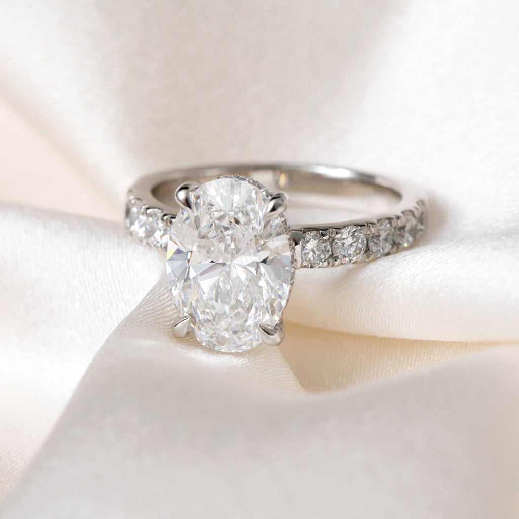 An oval-cut lab grown diamond engagement ring with a platinum band set with round brilliant cut diamonds, resting on a soft white satin fabric.