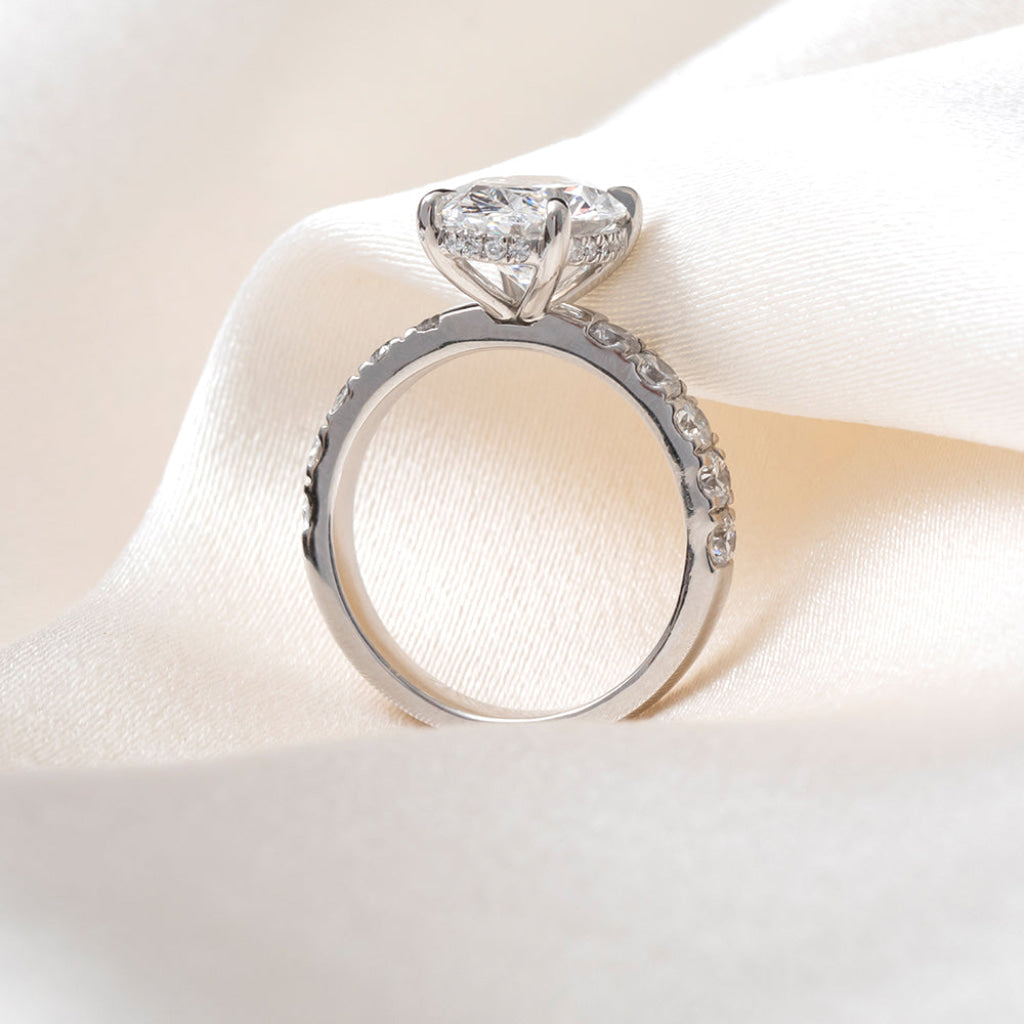 Side profile of an oval-cut diamond engagement ring with ap platinum band set with round brilliant cut diamonds, resting on a soft white satin fabric.