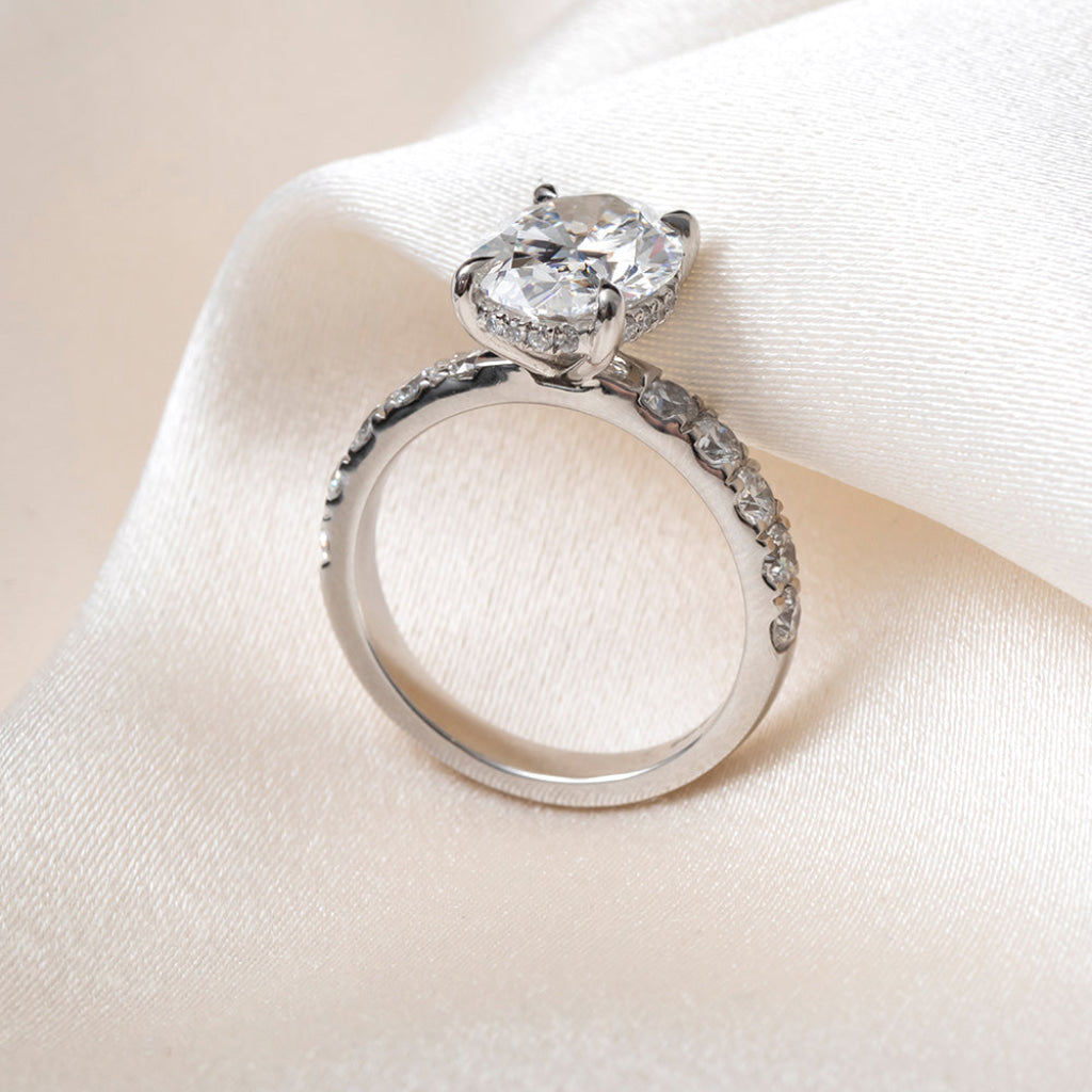 Side profile of an oval-cut diamond engagement ring with ap platinum band set with round brilliant cut diamonds, resting on a soft white satin fabric.