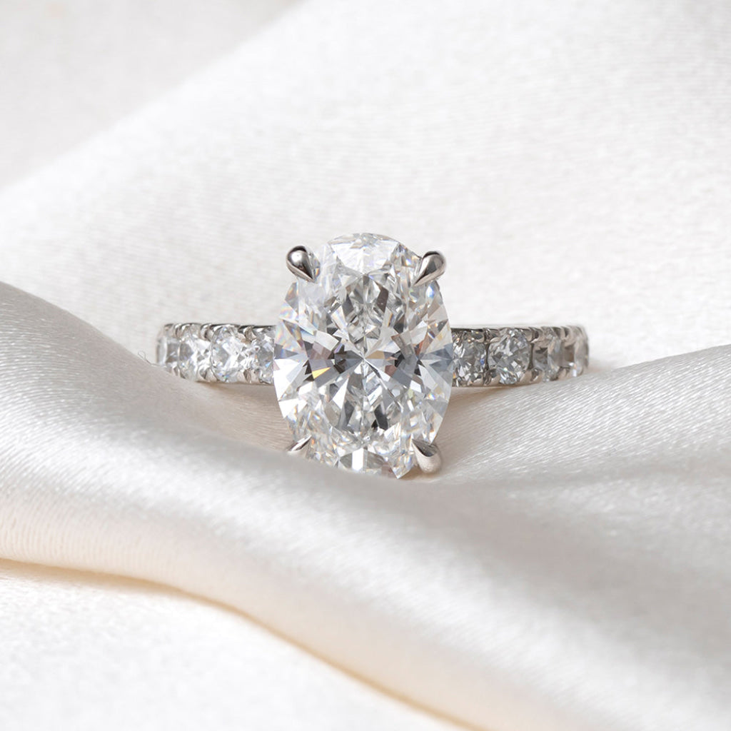 An oval-cut lab-grown diamond engagement ring with a platinum band set with round brilliant cut diamonds, resting on a soft white satin fabric.