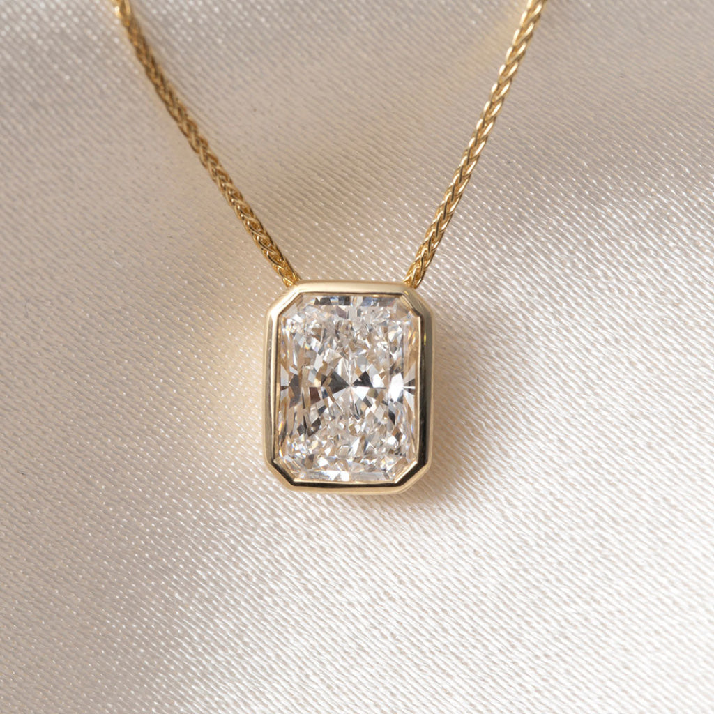 A yellow gold necklace featuring a radiant cut diamond pendant in a gold bezel setting, resting on a textured white satin fabric background.