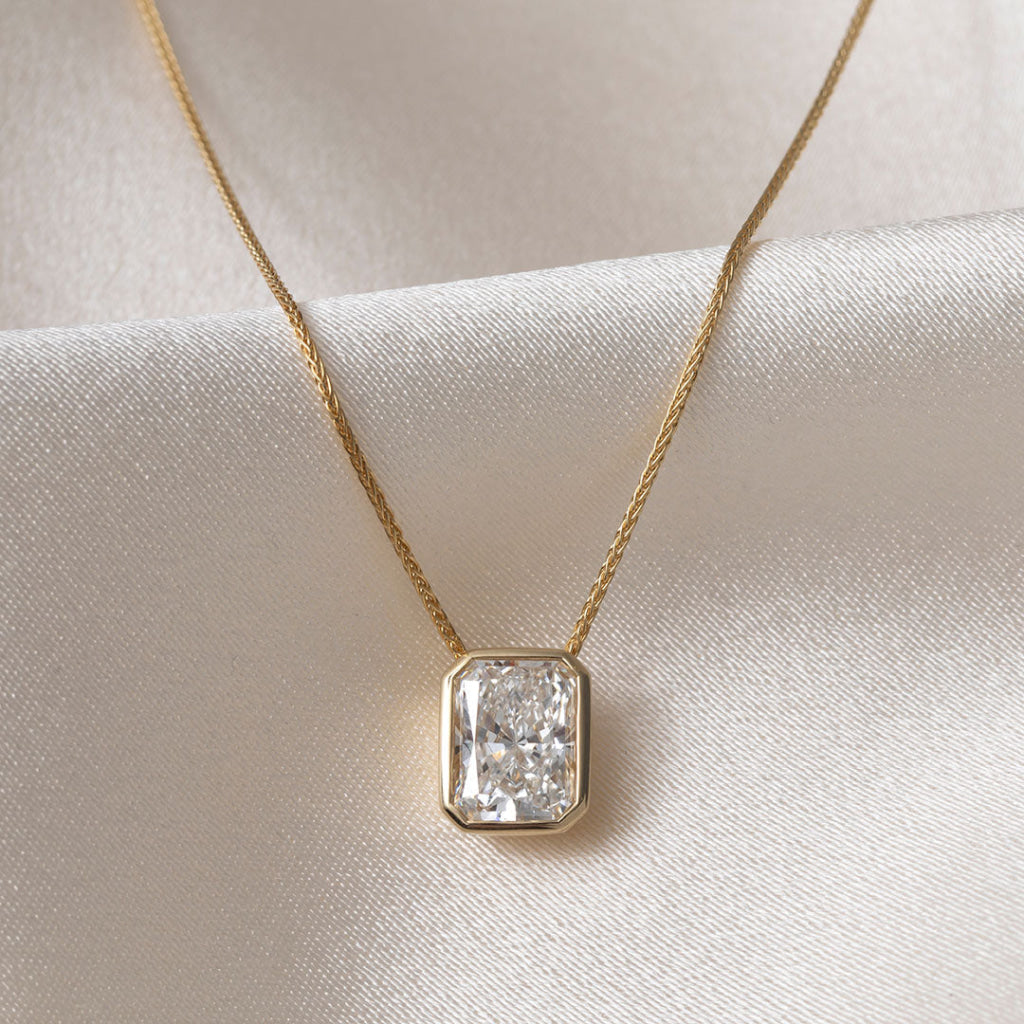 A yellow gold necklace featuring a radiant cut diamond pendant in a gold bezel setting, resting on a textured white satin fabric background.