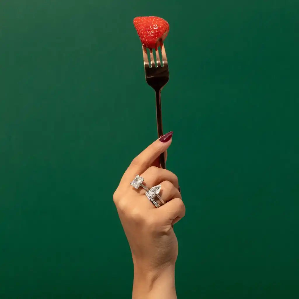 Fresh red strawberry perched on a silver fork held by a manicured hand wearing rings.