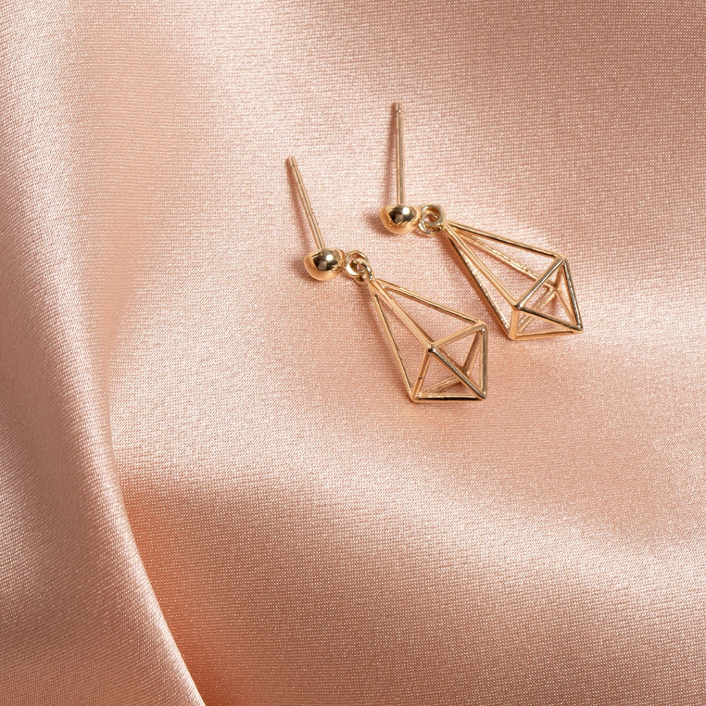 Gold geometric earrings with a shiny spherical stud on top and an open triangular prism-shaped dangle, casting a soft shadow on a cloth background