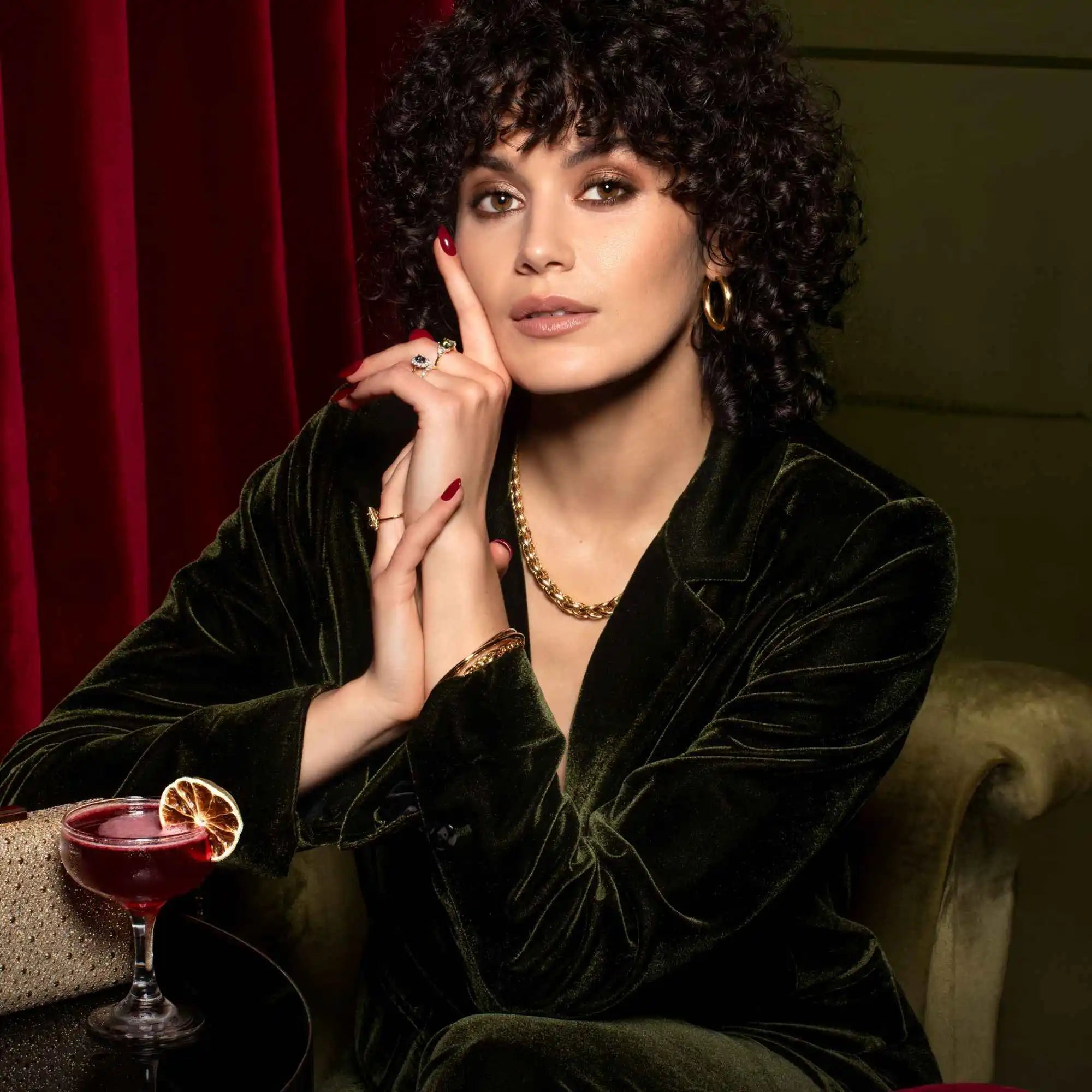 A glamorous portrait featuring curly dark hair, a green velvet blazer, and a red cocktail.