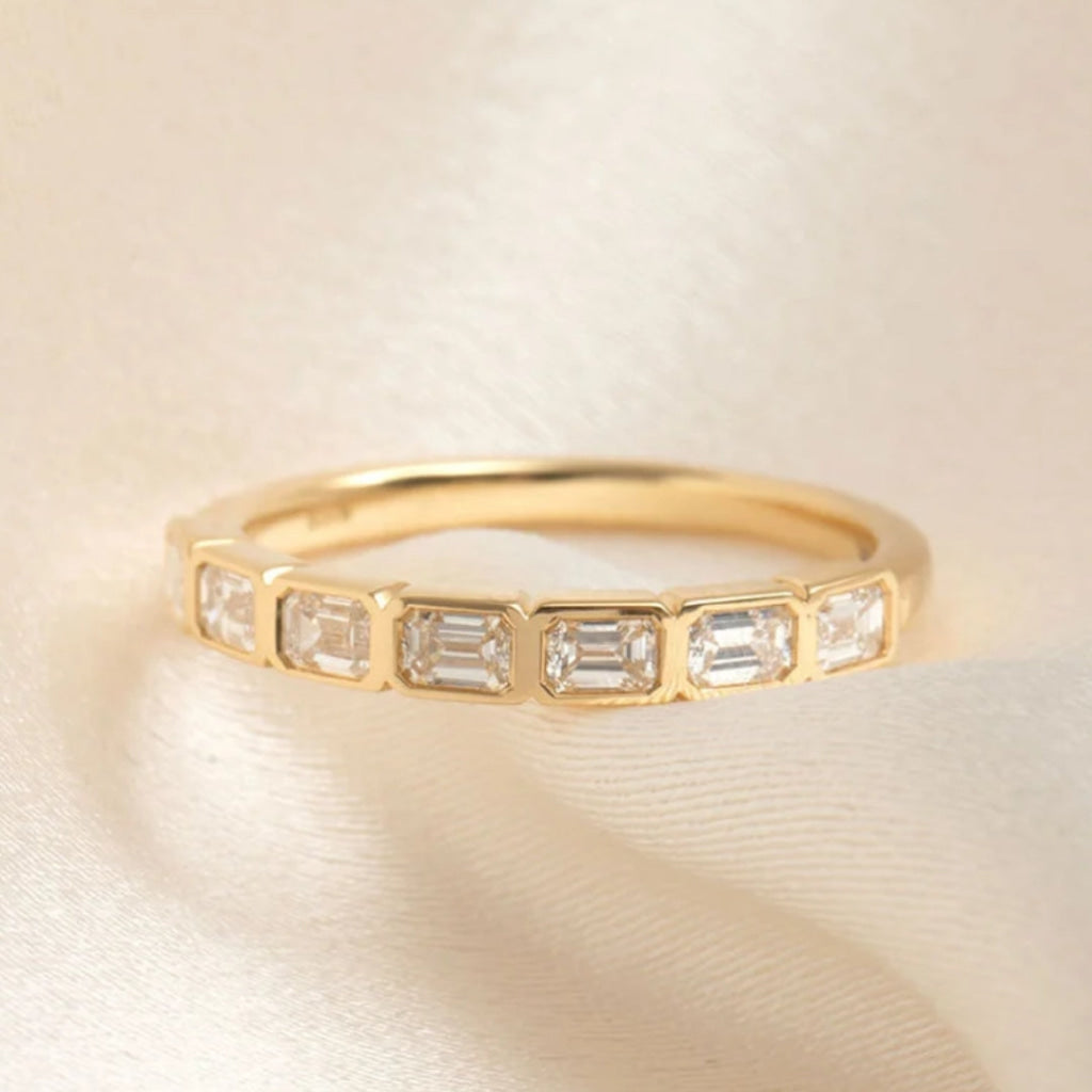 A yellow gold eternity ring featuring a row of baguette-cut clear gemstones, displayed on a soft, draped cream satin fabric.