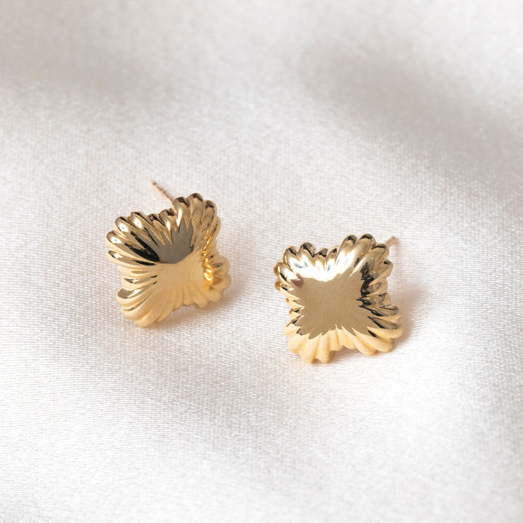 A pair of gold flower-shaped stud earrings resting on a textured white fabric background.