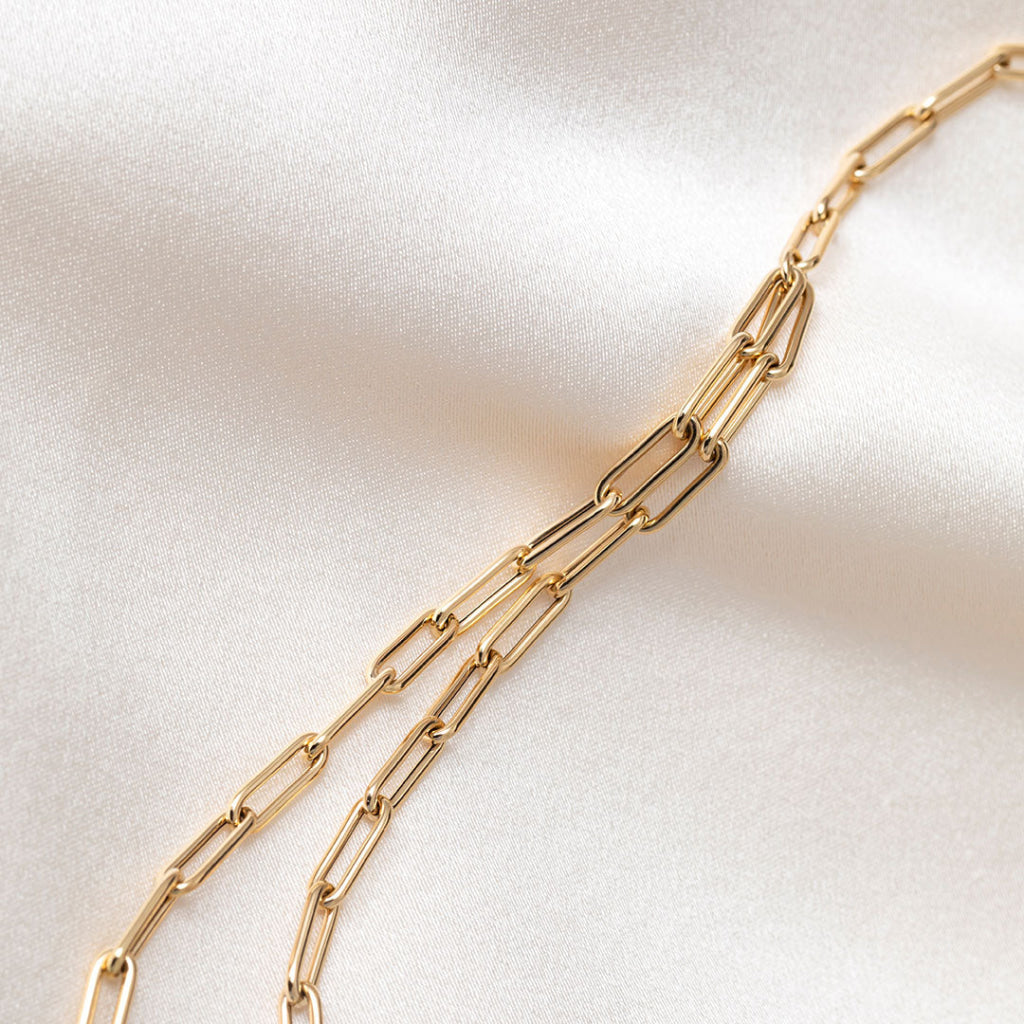 A gold paperclip chain necklace draped on a soft, white satin fabric background.