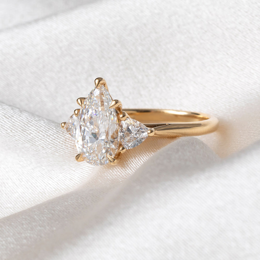 A yellow gold ring featuring a pear-cut center stone flanked by two trillion cut lab grown diamonds, displayed on a soft white satin fabric background.