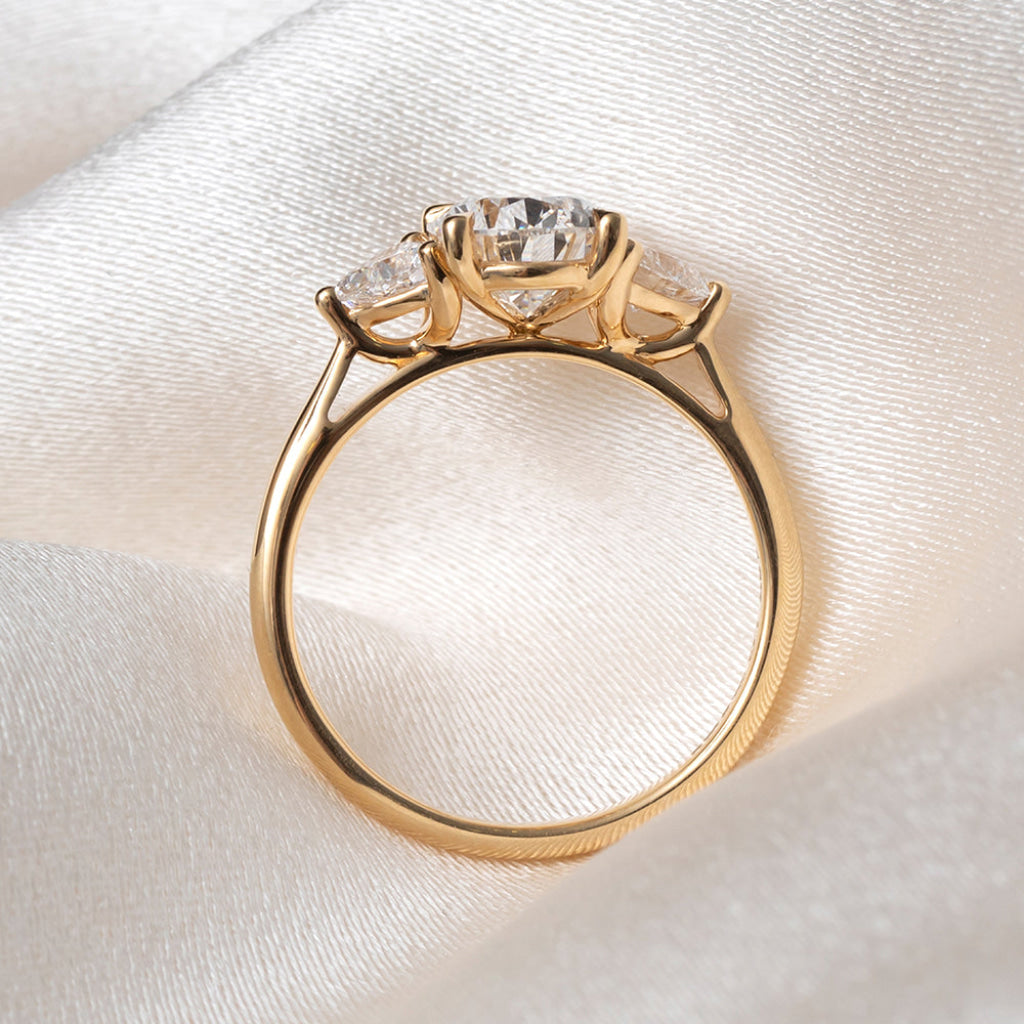 The side profile of a yellow gold ring featuring a pear-cut center stone flanked by two trillion cut lab grown diamonds, displayed on a soft white satin fabric background.
