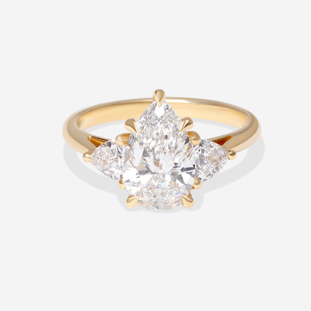 A yellow gold ring featuring a pear-cut center stone flanked by two trillion cut lab grown diamonds, displayed on a soft white background.