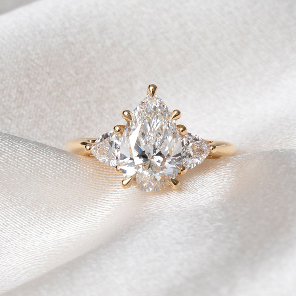 A yellow gold ring featuring a pear-cut center stone flanked by two trillion cut lab grown diamonds, displayed on a soft white satin fabric background.