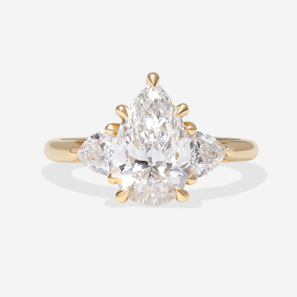 A yellow gold ring featuring a pear-cut center stone flanked by two trillion cut lab grown diamonds, displayed on a soft white background.