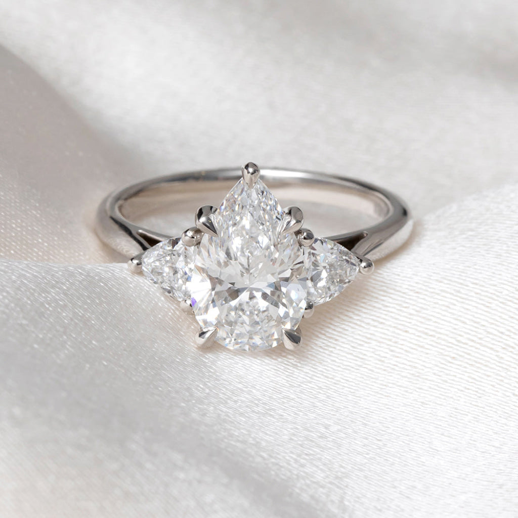 A platinum ring featuring a pear-cut center diamond flanked by two trillion cut lab-grown diamonds, displayed on a soft white fabric background.