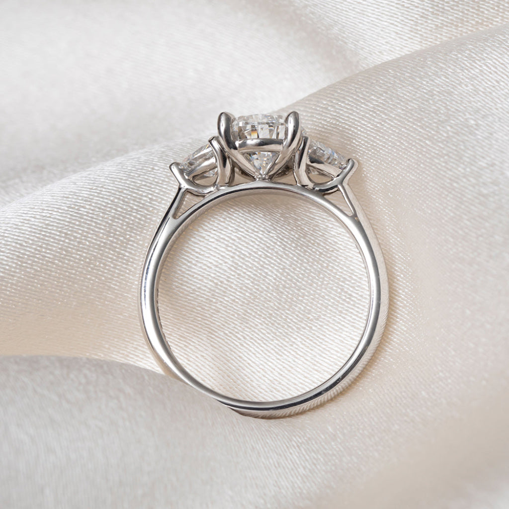 Side profile of a platinum ring featuring a pear-cut center diamond flanked by two trillion cut lab-grown diamonds, displayed on a soft white fabric background.