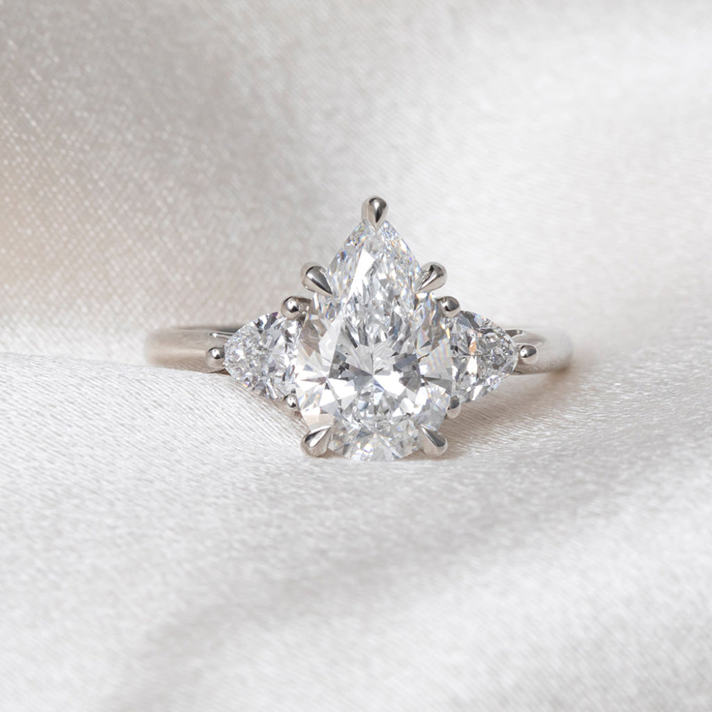 A platinum ring featuring a pear-cut center diamond flanked by two trillion cut lab-grown diamonds, displayed on a soft white fabric background.