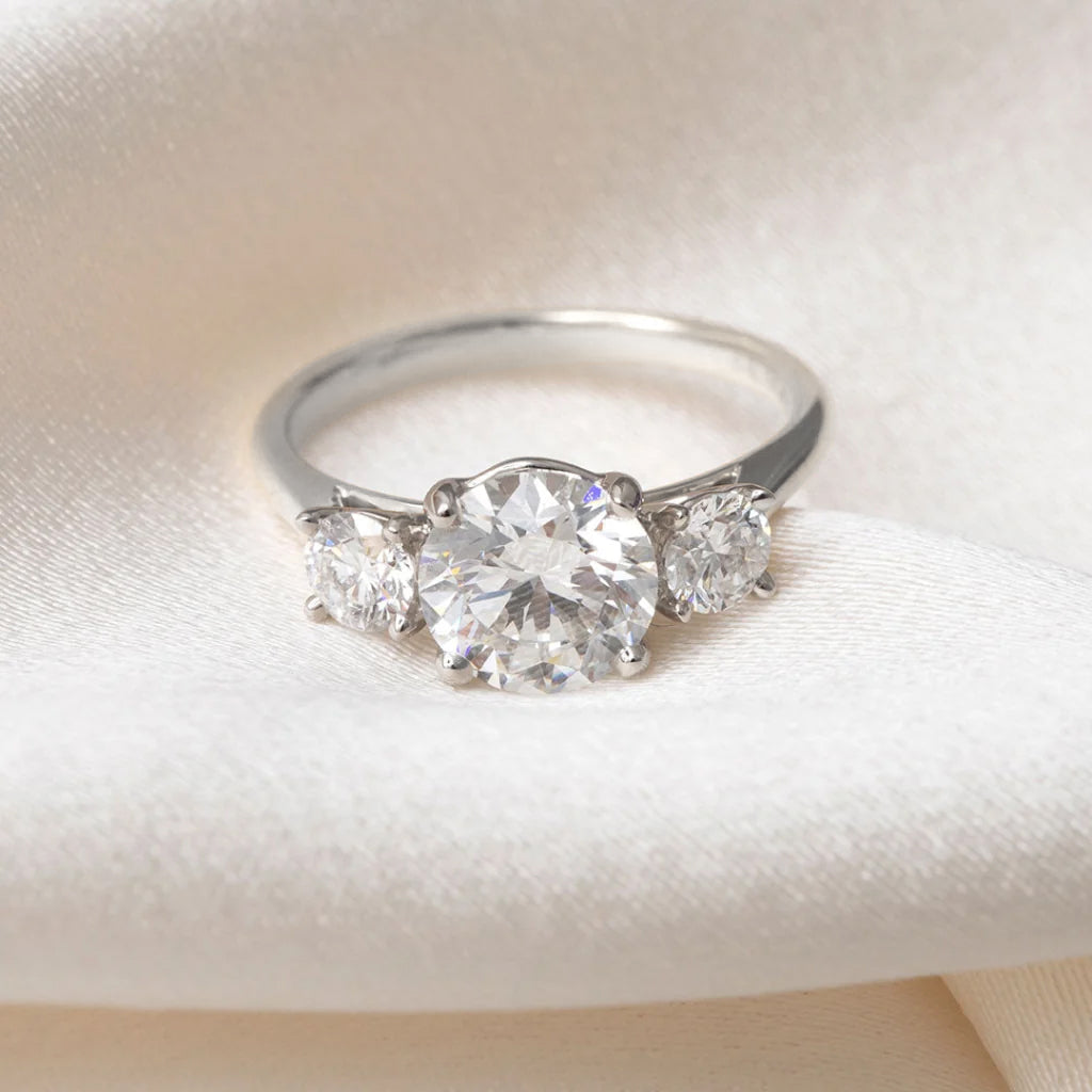 A platinum three-stone diamond ring with a large round center stone and two smaller side stones, displayed on a soft cream satin fabric.