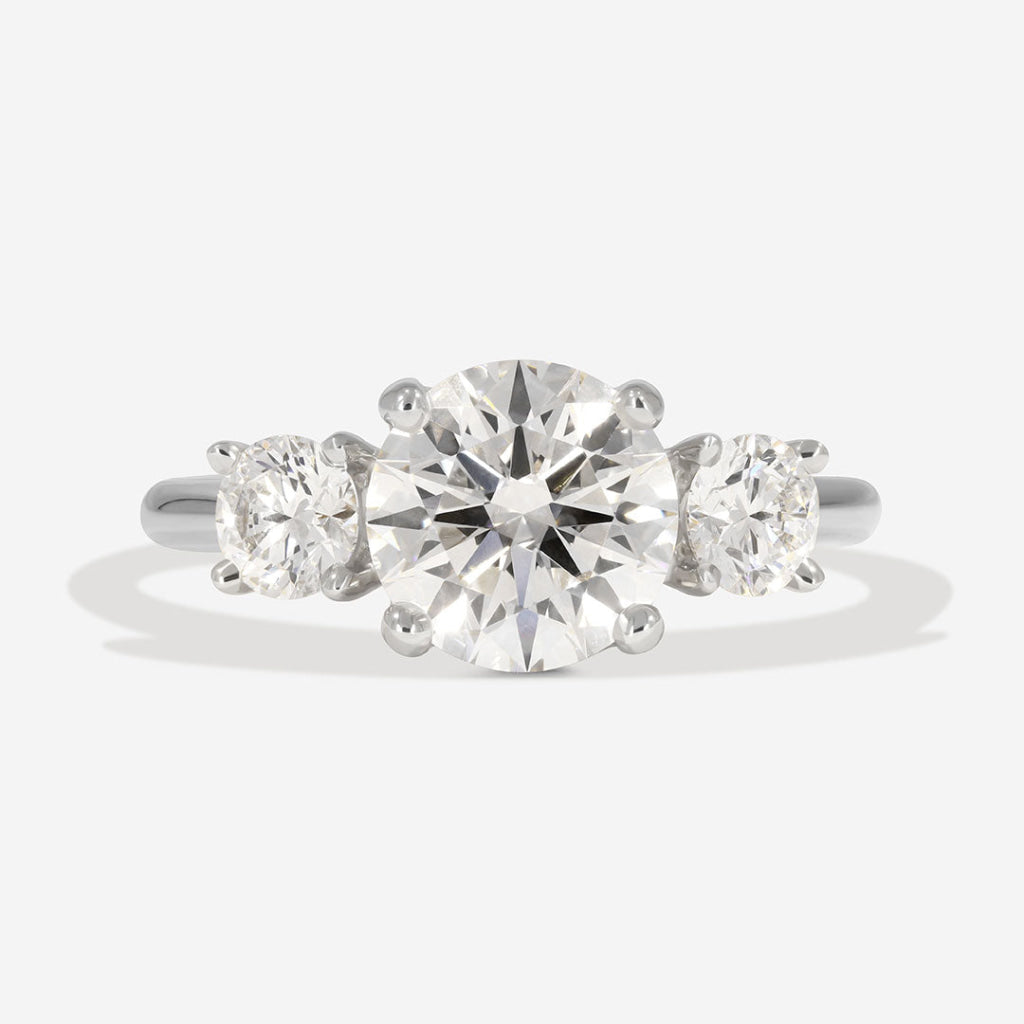 A three-stone diamond ring featuring a large round center diamond flanked by two smaller round side diamonds, set in a polished silver-colored metal band, shown on a light gray background.