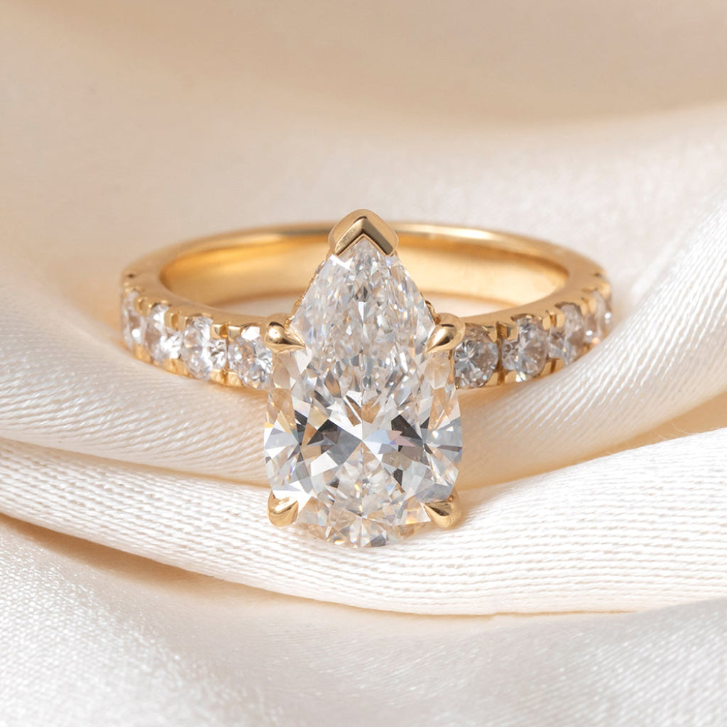 A yellow gold ring featuring a large pear-cut clear center stone and smaller round stones along the band, displayed on draped cream satin fabric.
