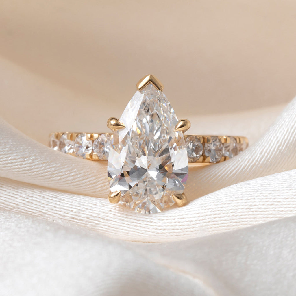 A gold ring featuring a large pear-cut clear center stone and smaller side stones, displayed on soft white satin fabric.