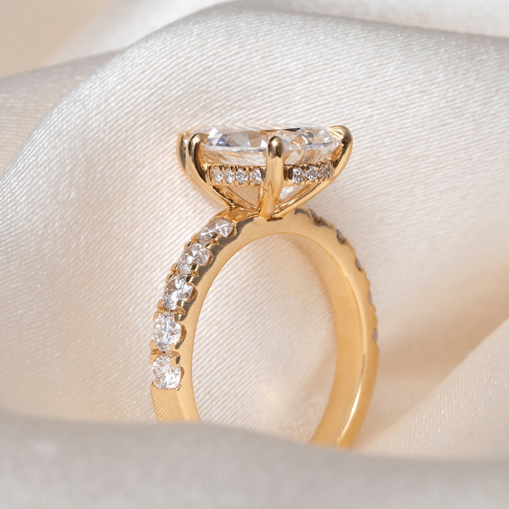 A side view of a yellow gold ring featuring a large pear-cut clear center stone and smaller round stones along the band, displayed on draped cream satin fabric.