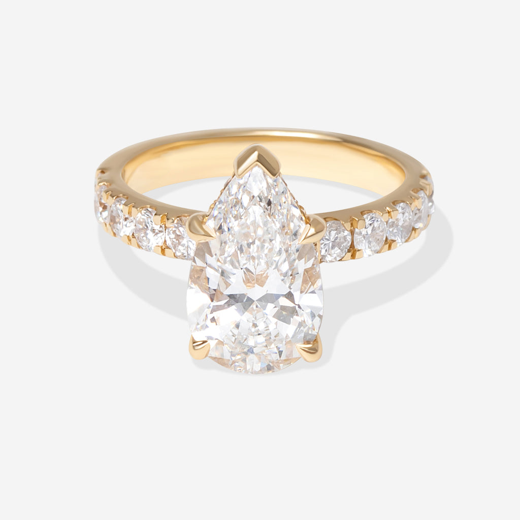 A yellow gold ring featuring a large pear-cut center diamond and smaller round diamonds along the shoulders, set against a light gray background.