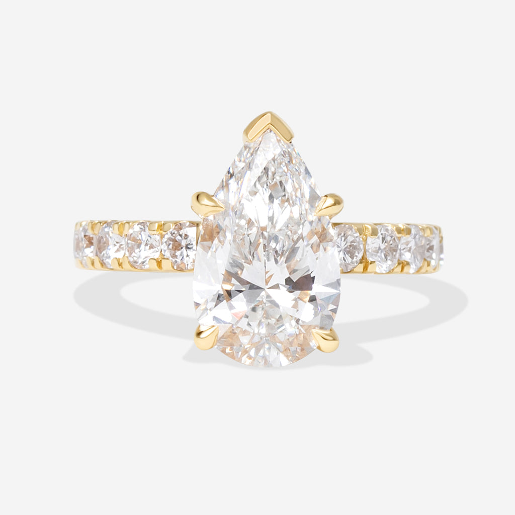 A pear-cut diamond engagement ring in yellow gold with a pavé band, set against a light gray background.