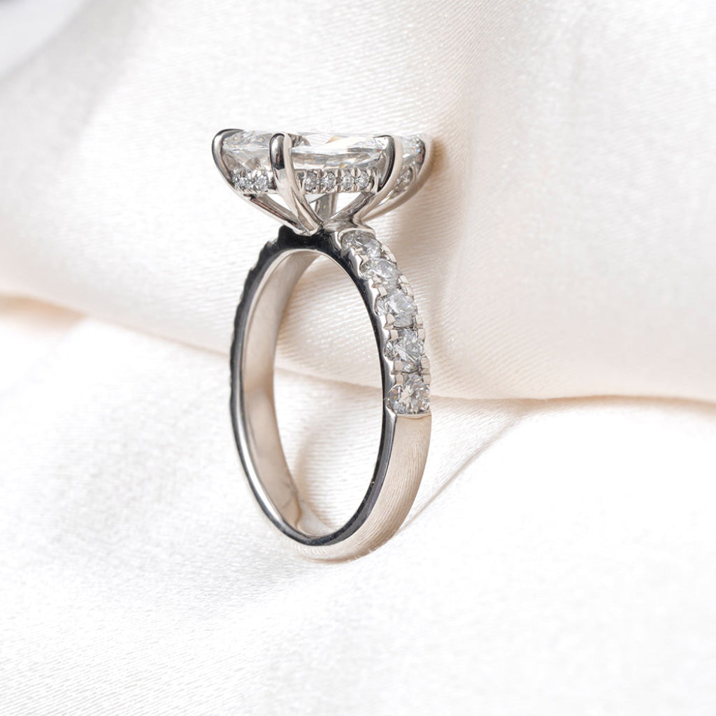 A platinum solitaire engagement ring with a pear cut center stone and pavé-set side stones, displayed on a soft white satin fabric background.