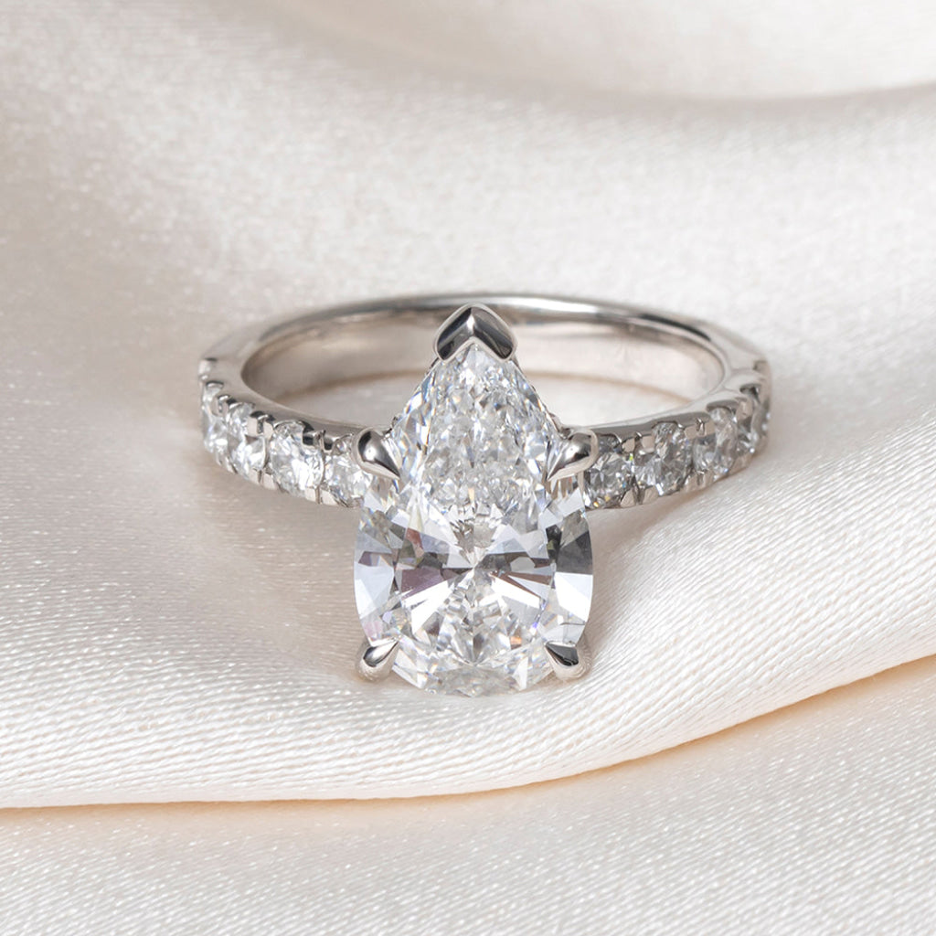 A pear-cut diamond engagement ring with a platinum band accented by pavé diamonds, resting on a draped cream satin fabric.