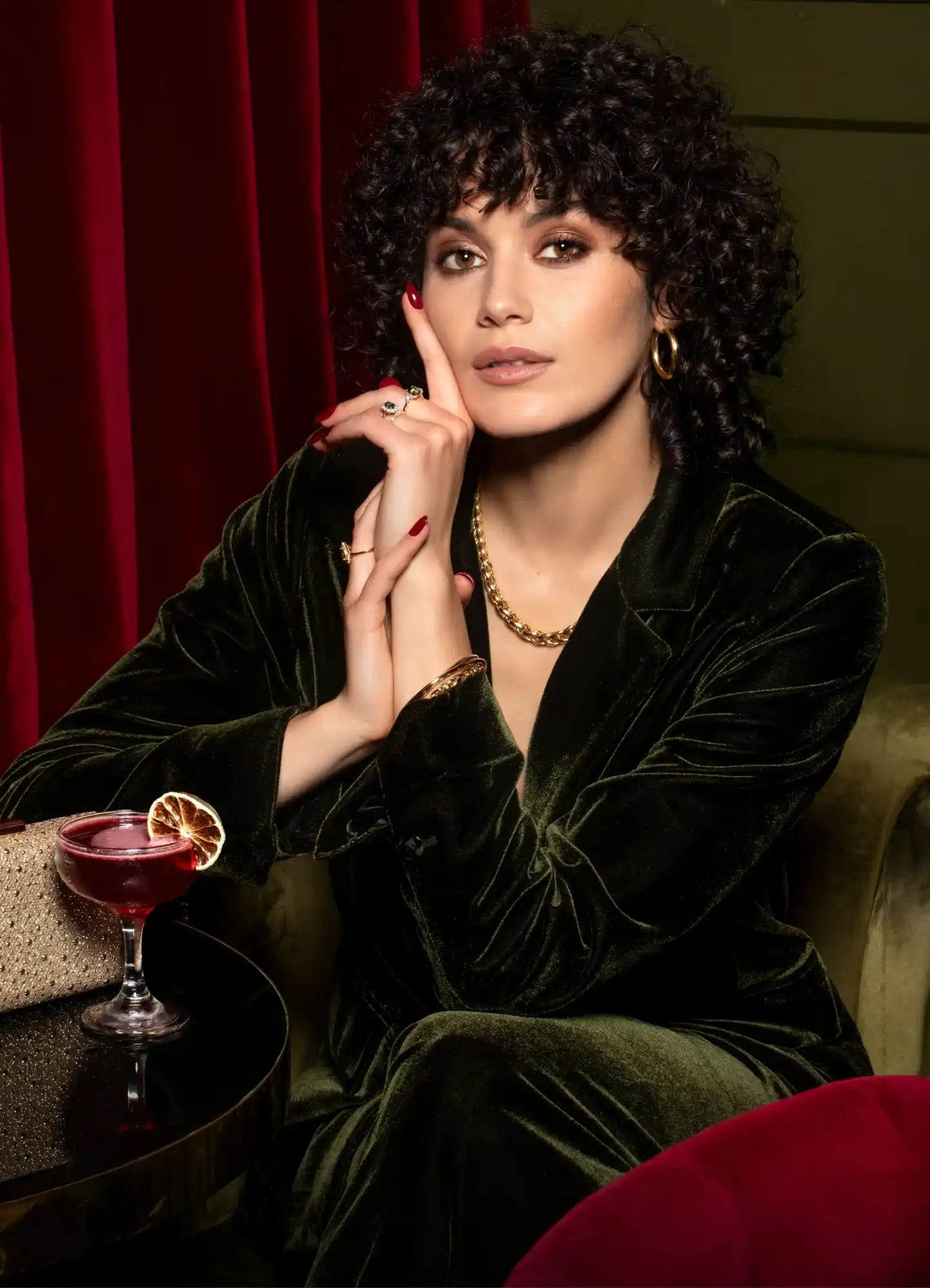 A person in a dark green velvet dress posing with a cocktail.