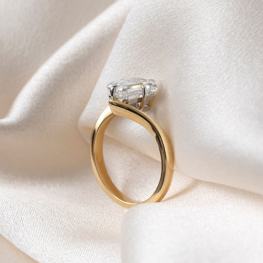 An emerald-cut diamond ring with a yellow gold band, resting on draped cream-colored satin fabric.