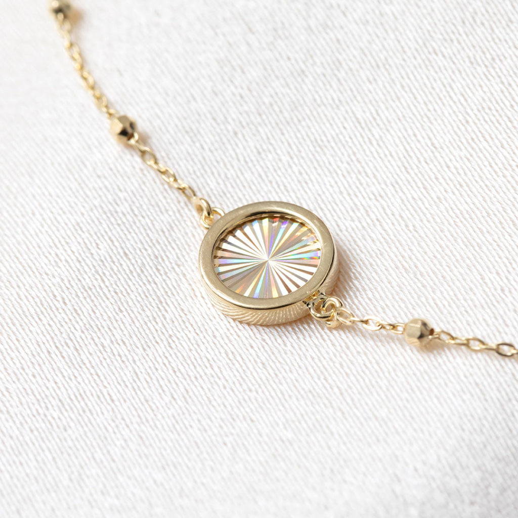 A delicate gold chain bracelet with small bead accents and a central round disc, displayed on a soft white fabric background.